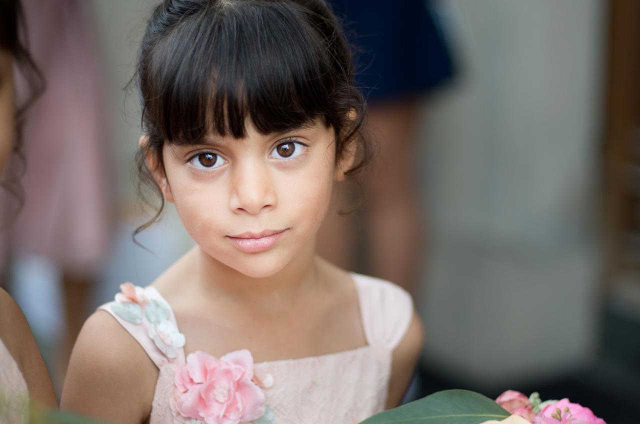 Portret dziewczynki w jasnoróżowej sukience z kwiatami, spoglądającej w obiektyw z delikatnym uśmiechem, na rozmytym tle.