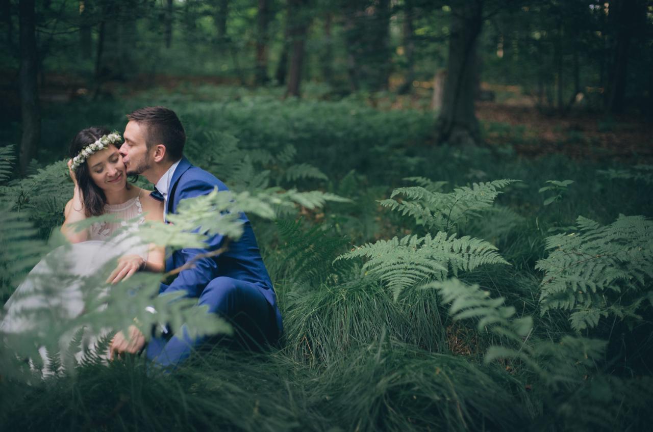 Panna młoda w wianku i pan młody w niebieskim garniturze siedzą w lesie, pan młody całuje pannę młodą w policzek, otoczeni paprociami.