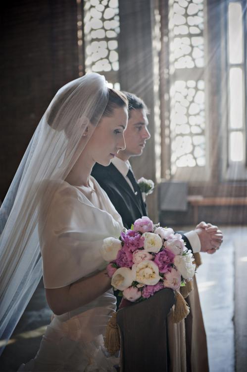 Panna młoda w welonie i bukietem piwonii oraz pan młody w ciemnym garniturze, oboje w kościele, światło słoneczne wpada przez okna.