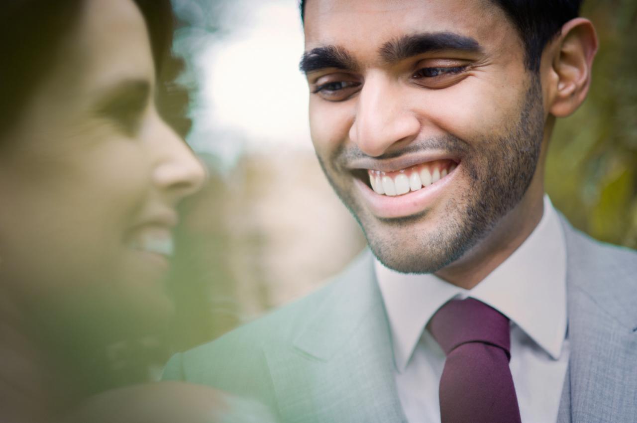 Uśmiechnięty pan młody w szarym garniturze i fioletowym krawacie, obok rozmazana sylwetka panny młodej.