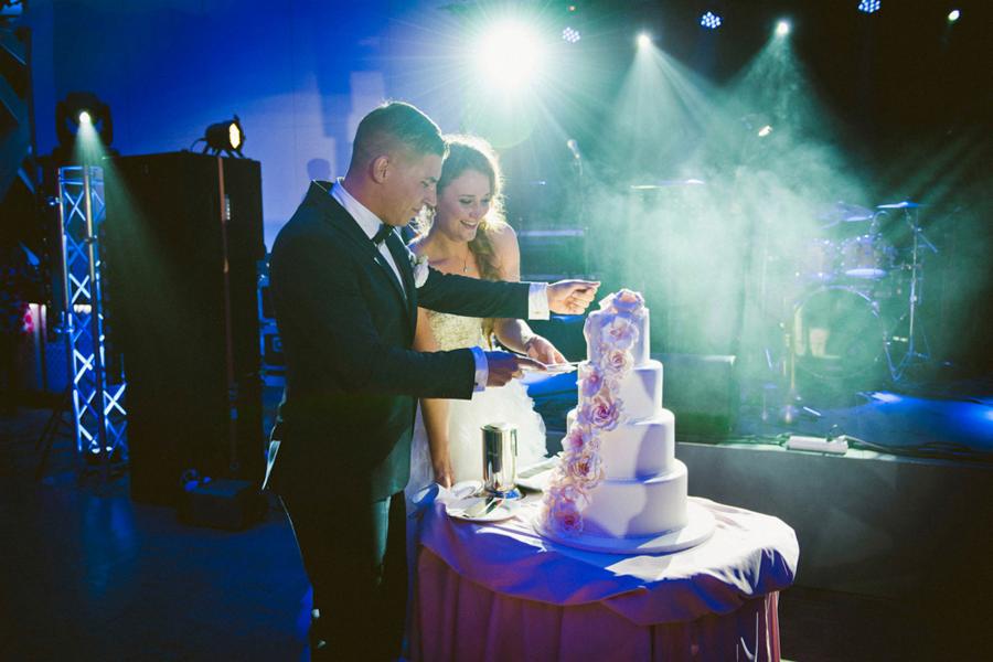 Para młoda kroi tort weselny na tle sceny z instrumentami, oświetlonej reflektorami.