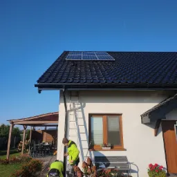 Montaż paneli fotowoltaicznych na dachu domu jednorodzinnego z czarną dachówką, dwóch instalatorów w jaskrawych kurtkach pracuje na wysokości, jeden na drabinie, drugi przy ziemi, błękitne niebo...