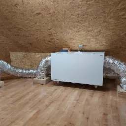 Biała centrala wentylacyjna z podłączonymi, srebrnymi przewodami wentylacyjnymi na drewnianych podkładkach, umieszczona na poddaszu z drewnianą boazerią.