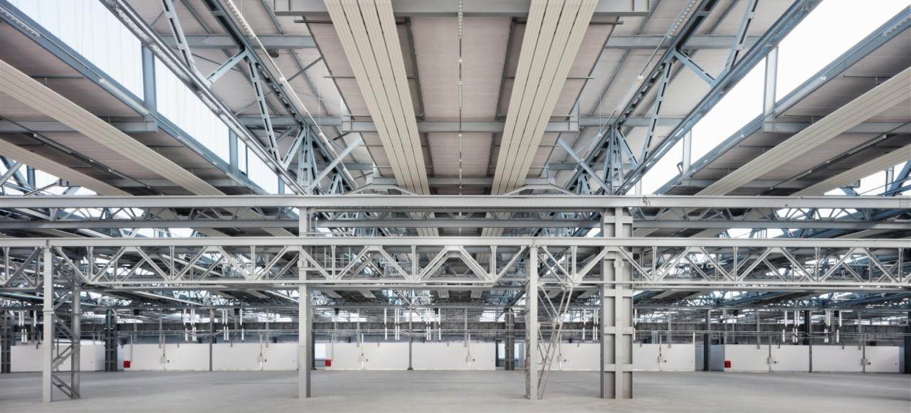Wnętrze hali przemysłowej z widoczną stalową konstrukcją nośną, betonową posadzką i białymi ścianami działowymi.