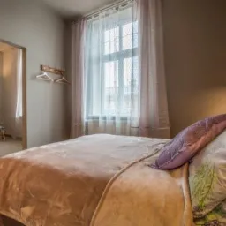 Beżowa sypialnia z łóżkiem z pluszowym kocem, dekoracyjną poduszką i lampą wiszącą, widok na pokój obok z krzesłem i kinkietem