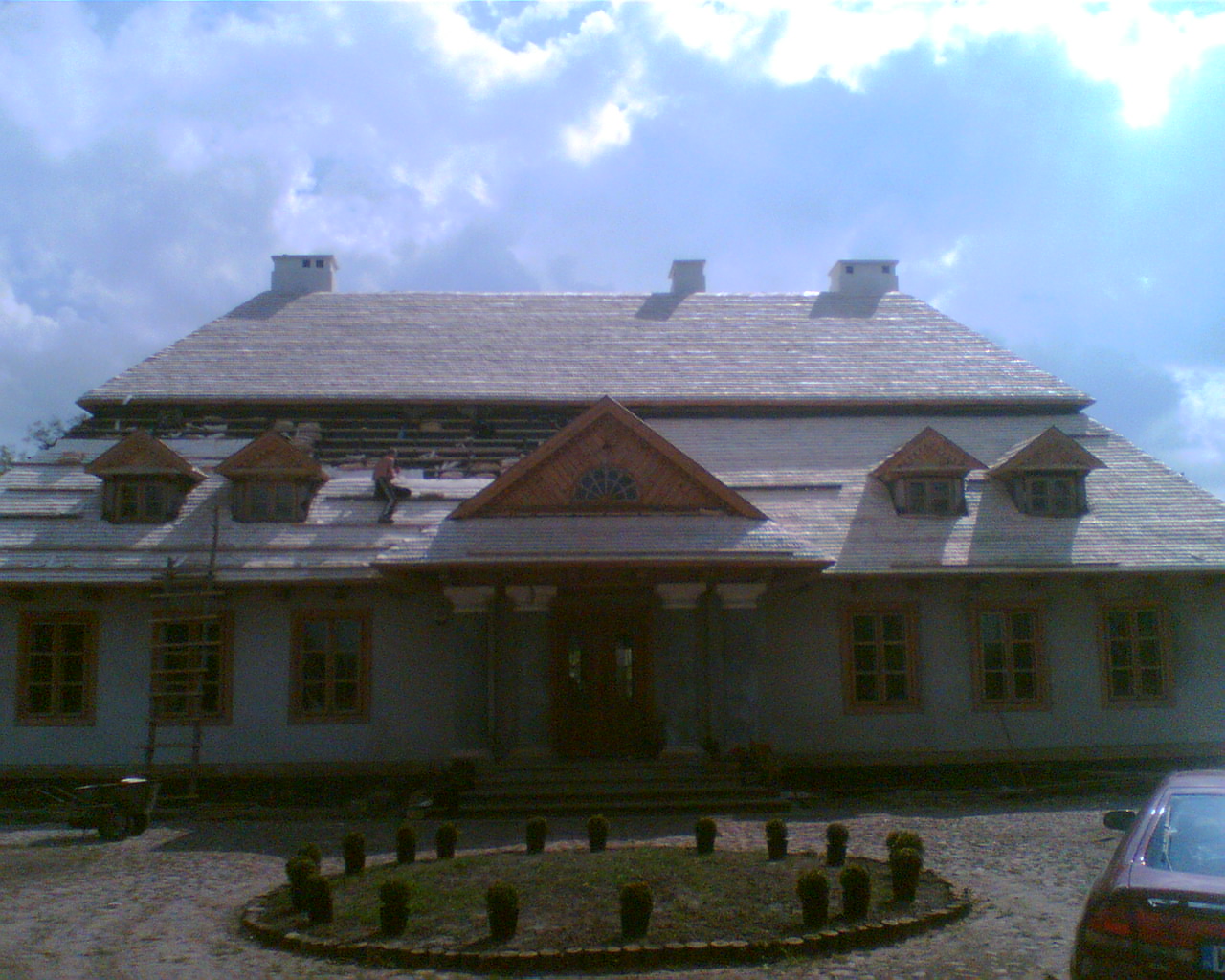 Dworek z drewnianym dachem w trakcie naprawy, widoczny dekarz na dachu i częściowo zdjęte poszycie, na pierwszym planie ozdobny klomb.