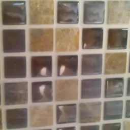 Mozaika ścienna z kwadratowych płytek w odcieniach brązu, beżu i szarości, z błyszczącą powierzchnią i jasną fugą.