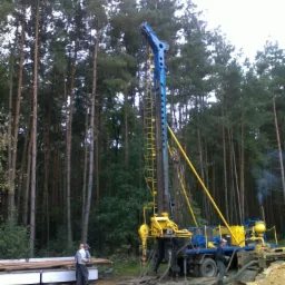 Wysoka, niebiesko-żółta wiertnica na podwoziu ciężarówki, ustawiona na skraju lasu sosnowego. Obok leżą rury w białej obudowie, a dwóch mężczyzn obserwuje pracę maszyny. Z rur wiertniczych wydobywa...