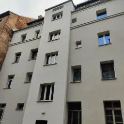 Odnowiona fasada budynku wielorodzinnego z widocznymi oknami, świeżo pomalowana na jasny kolor, z numerem 3 nad wejściem, sąsiadująca z budynkiem z czerwonej cegły.