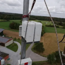 Widok z góry na zamontowane na metalowym słupie urządzenia telekomunikacyjne, zabezpieczone liną, z widocznym budynkiem z panelami słonecznymi i polami w tle.