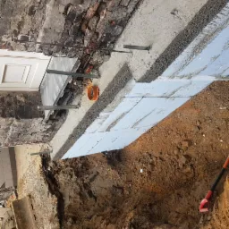 Wykop pod fundamenty z izolacją termiczną i zbrojeniem przy ceglanej ścianie budynku, widoczna rura kanalizacyjna i łopata.