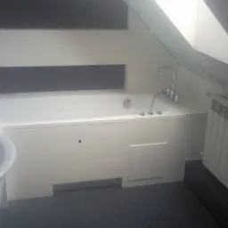 Biała wanna zabudowana w łazience na poddaszu, bateria wannowa z chromowanymi kurkami, grzejnik żeberkowy pod skosem, fragment umywalki z widocznym rantem, płytki w odcieniach bieli, szarości...