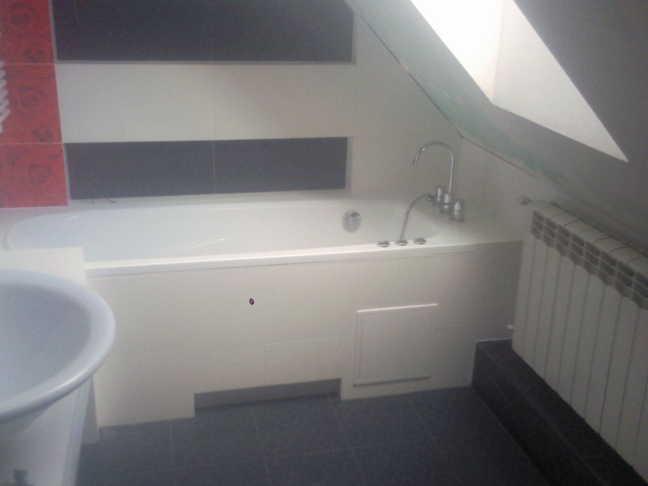 Biała wanna zabudowana w łazience na poddaszu, bateria wannowa z chromowanymi kurkami, grzejnik żeberkowy pod skosem, fragment umywalki z widocznym rantem, płytki w odcieniach bieli, szarości...