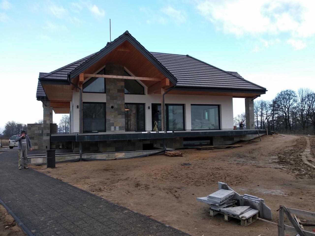 Nowoczesny dom jednorodzinny z elementami kamiennymi w trakcie wykańczania elewacji, widoczny pracownik w kombinezonie i ułożona ścieżka z kostki brukowej.