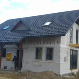 Dom w trakcie budowy z ciemnym dachem z dachówki modułowej i oknami dachowymi, widoczny pracownik przy wejściu.