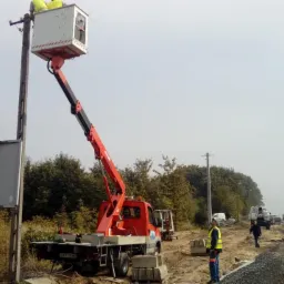 Prace przy słupie energetycznym z użyciem podnośnika koszowego, widoczny pracownik w koszu oraz drugi na ziemi w kamizelce odblaskowej, w tle inne pojazdy i drzewa.