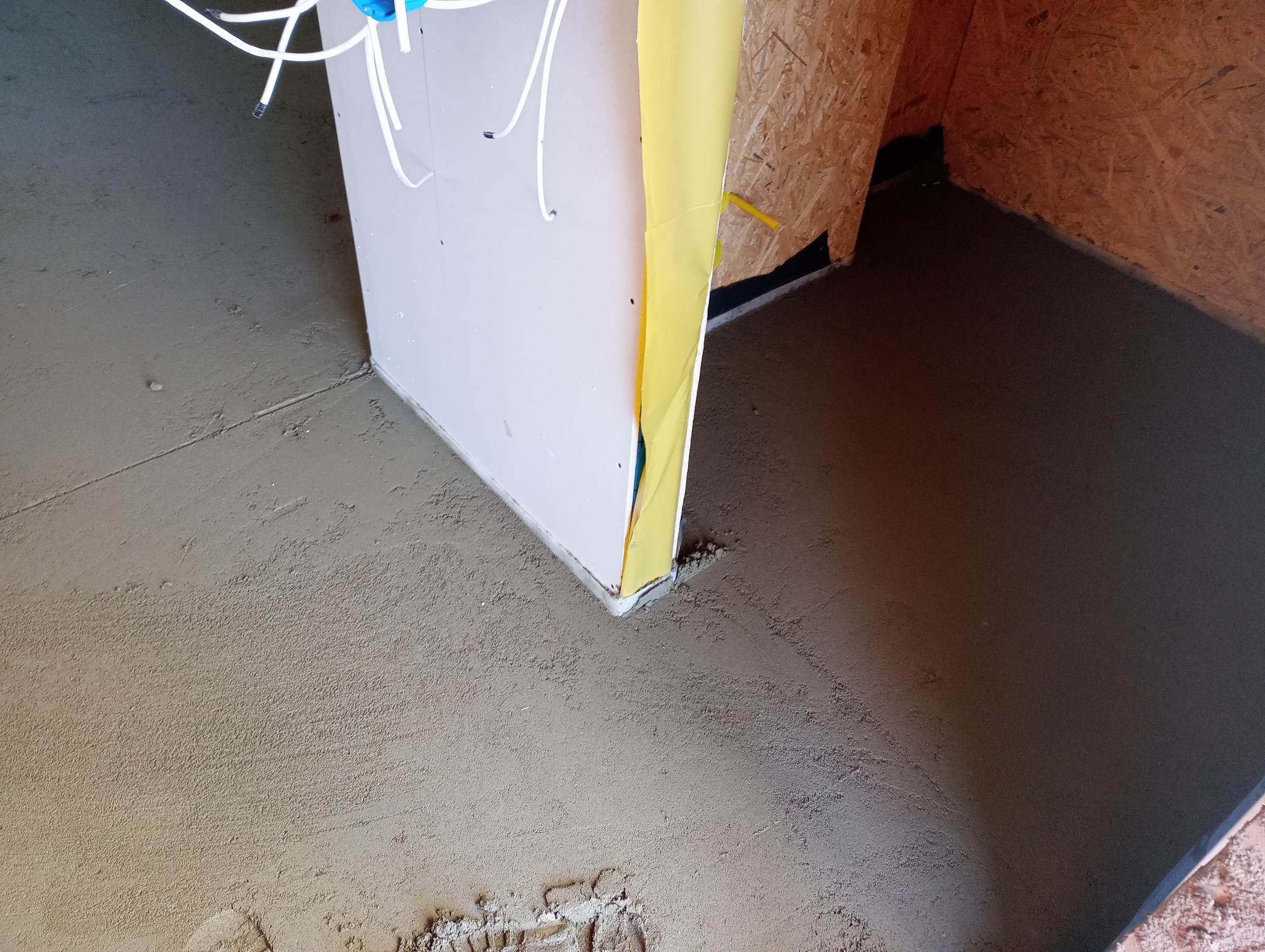 Świeżo wylana, wilgotna posadzka jastrychowa przy ścianie z widoczną folią ochronną i wystającymi przewodami elektrycznymi.