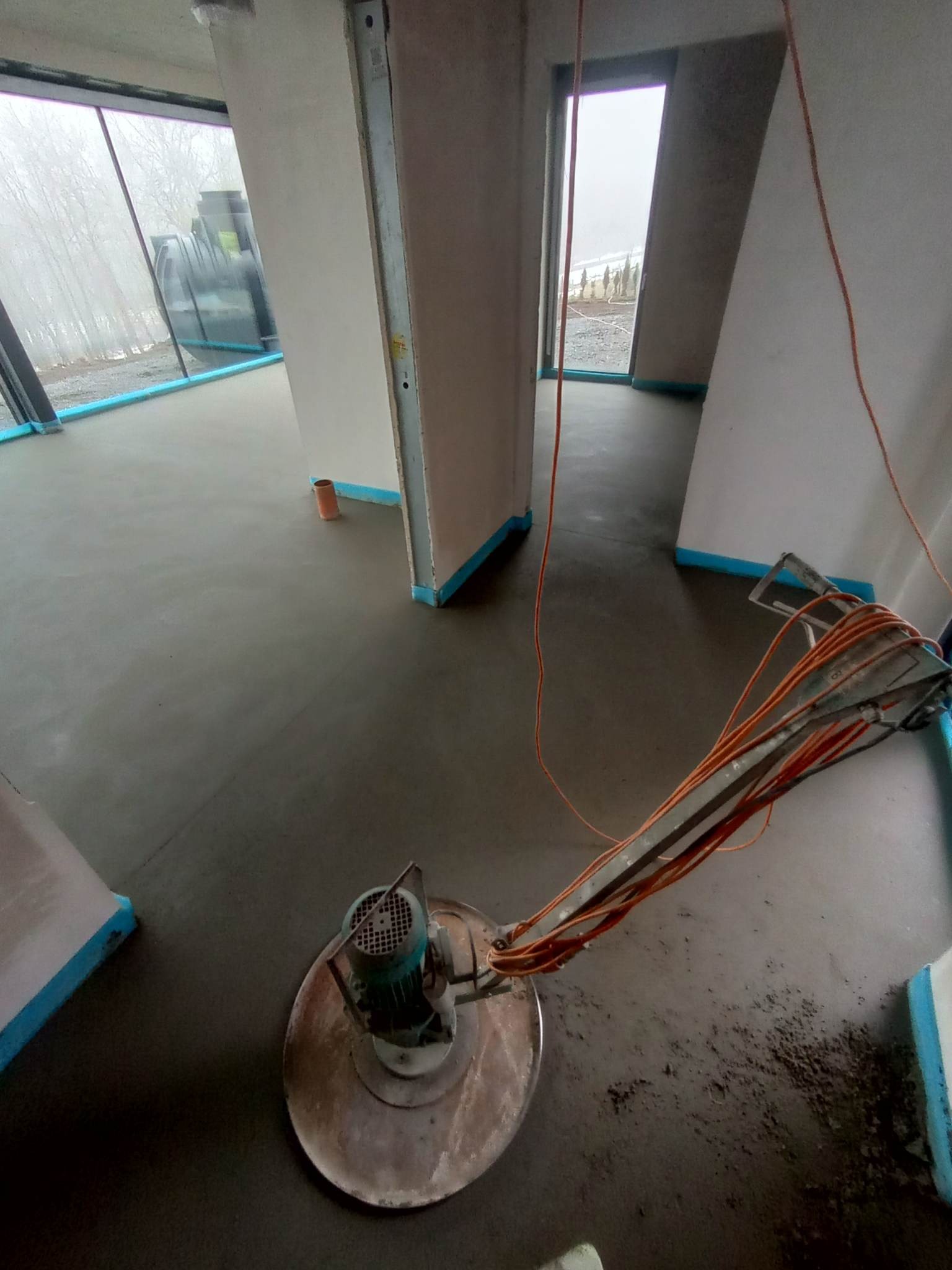 Świeżo wylana, gładka posadzka z jastrychu cementowego w trakcie obróbki mechanicznej za pomocą szlifierki talerzowej; widoczne zabezpieczenia folią przy ścianach i oknach.