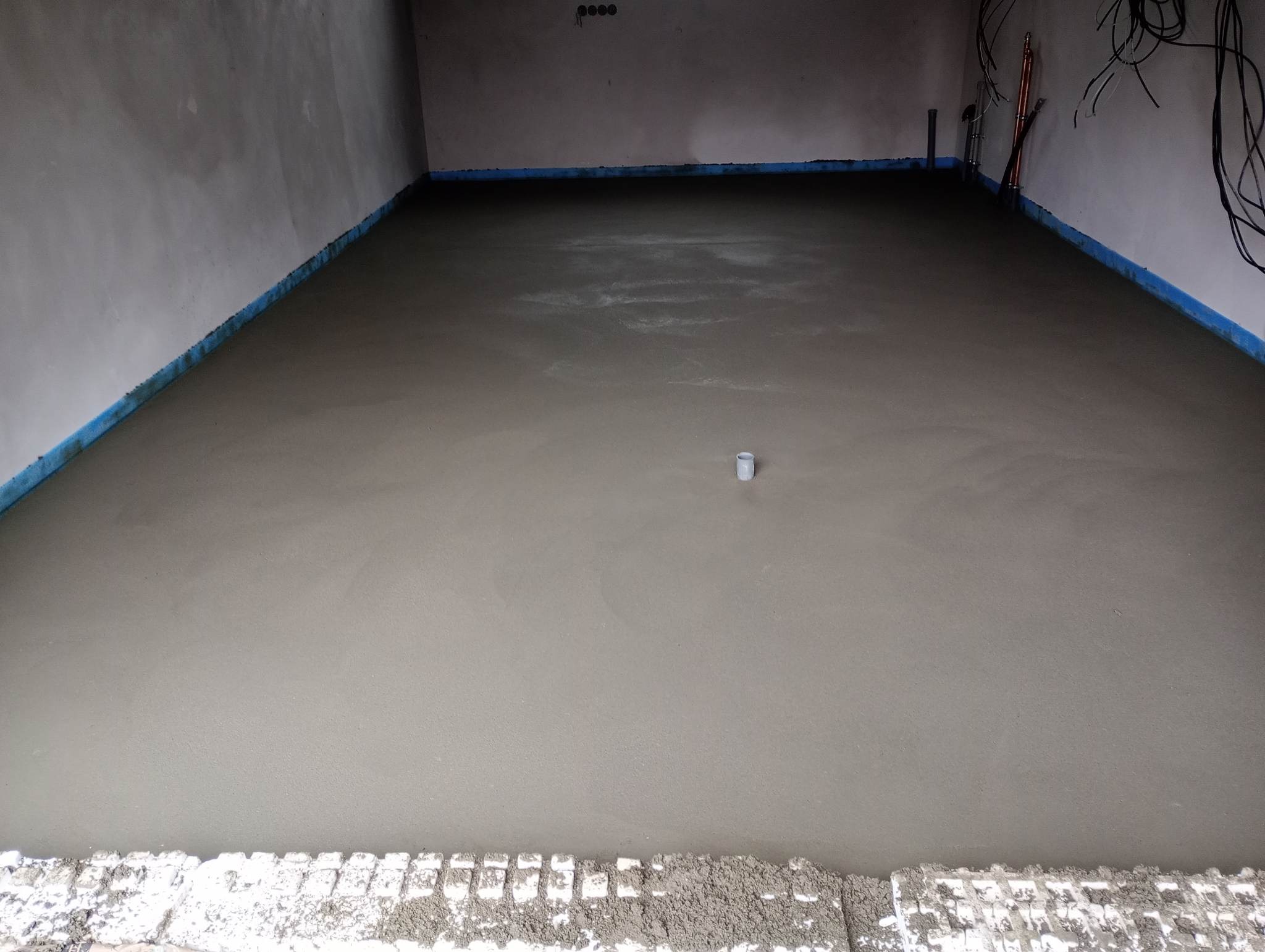 Świeżo wylana, gładka posadzka jastrychowa w pomieszczeniu z zabezpieczonymi ścianami folią i widocznymi przewodami elektrycznymi.