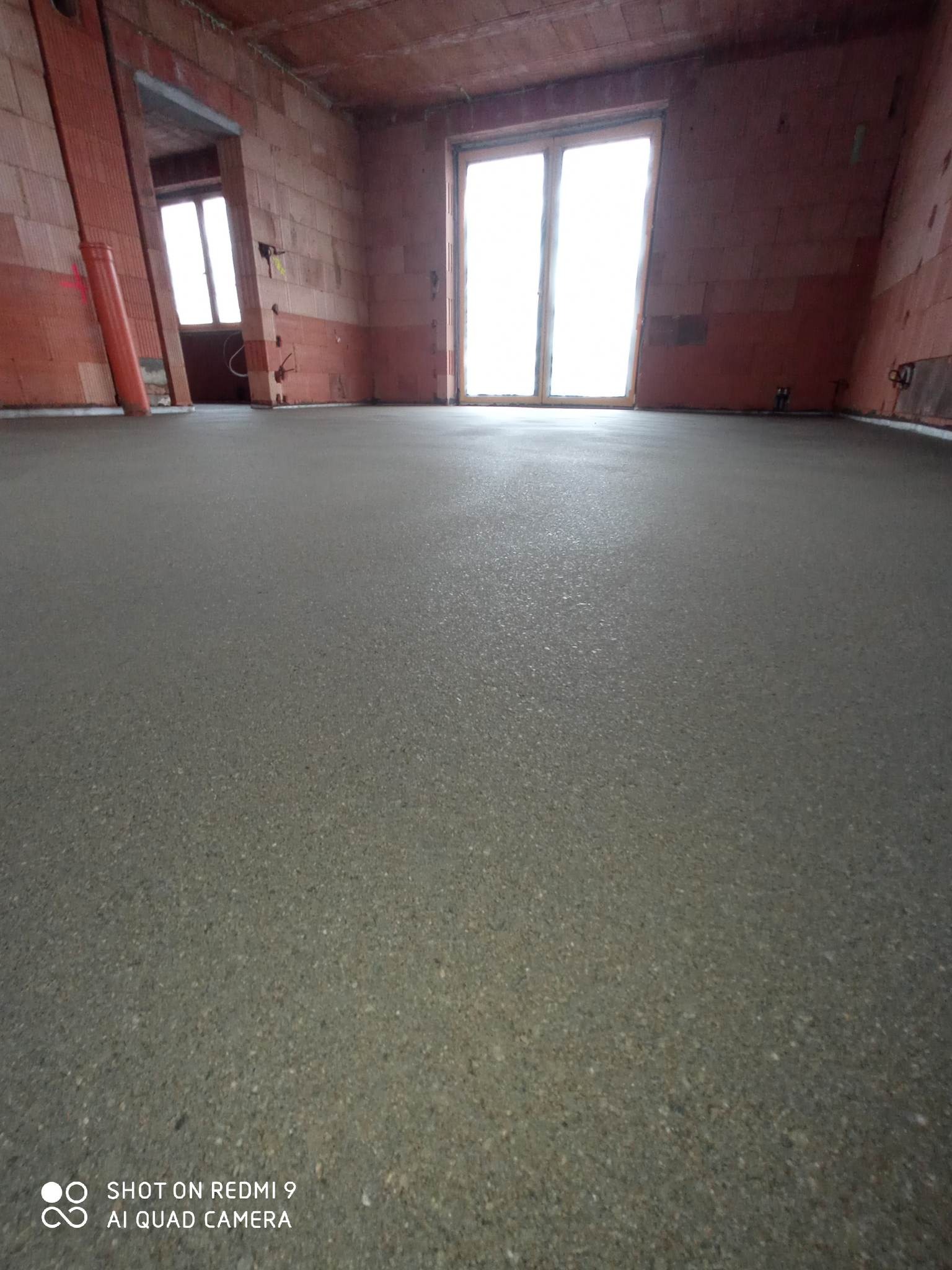 Świeżo wylana, gładka posadzka w surowym wnętrzu z czerwonej cegły, widoczne okno i fragment rury przy ścianie.