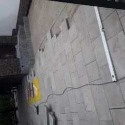 Układanie prostokątnej kostki granitowej w kolorze szarym, widoczna poziomica, fragment budynku z cegły i żółta mata ochronna.