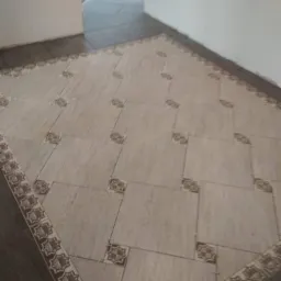 Ułożona podłoga z płytek ceramicznych w odcieniach beżu i brązu, z dekoracyjnym obramowaniem i krzyżykami dystansowymi między płytkami, widoczne przejście do innego pomieszczenia z ciemniejszą...