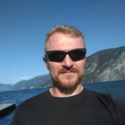 Mężczyzna w okularach przeciwsłonecznych robiący selfie na tle fiordu z górami i błękitnym niebem. Widoczny fragment pomostu i inni ludzie.