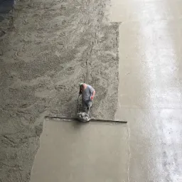 Pracownik wyrównuje wibrołatą świeżo wylaną, szarą posadzkę w hali przemysłowej, widok z góry na fragment surowego betonu i gładką powierzchnię.