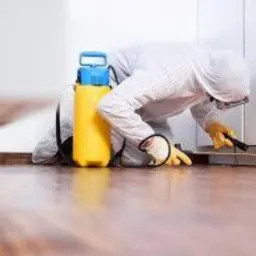 Osoba w kombinezonie ochronnym i okularach, z żółtym opryskiwaczem obok, aplikuje preparat w szczelinę między podłogą a ścianą przy użyciu małego pędzelka.