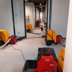 DRY-TECH OSUSZANIE BUDYNKÓW - Korytarz biurowy z rozstawionymi, pracującymi osuszaczami powietrza Wilms w kolorach żółto-czerwonym. Widoczne przewody i detale urządzeń. Perspektywa w głąb korytarza.