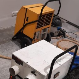 DRY-TECH OSUSZANIE BUDYNKÓW - Dwa przemysłowe urządzenia do osuszania, żółte i białe, z czarnymi uchwytami, stoją na szarej podłodze w pomieszczeniu. W tle widoczny odkurzacz i fragment ściany.