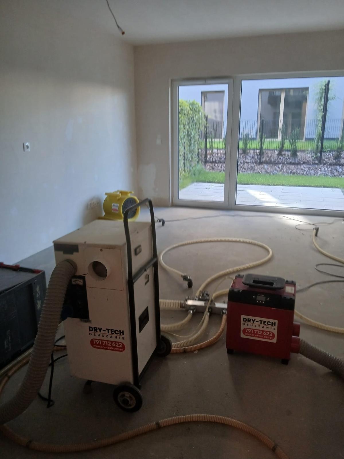 Dwa urządzenia do osuszania, białe i czerwone, z podłączonymi wężami, stoją w pustym, jasnym pomieszczeniu z dużym oknem. Widoczne logo firmy na urządzeniach.