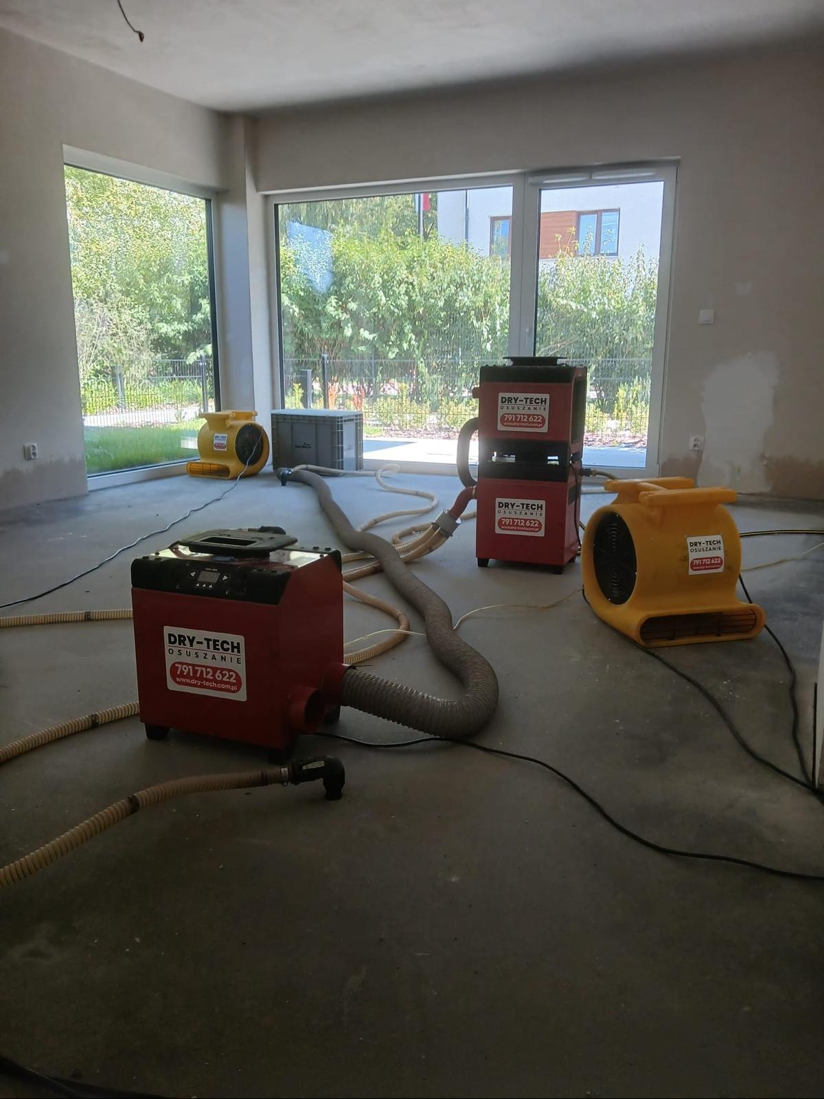 Wnętrze w trakcie prac. Widoczne urządzenia do osuszania, wentylatory i węże. Duże okna z widokiem na zieleń. Szare ściany i podłoga w surowym stanie.