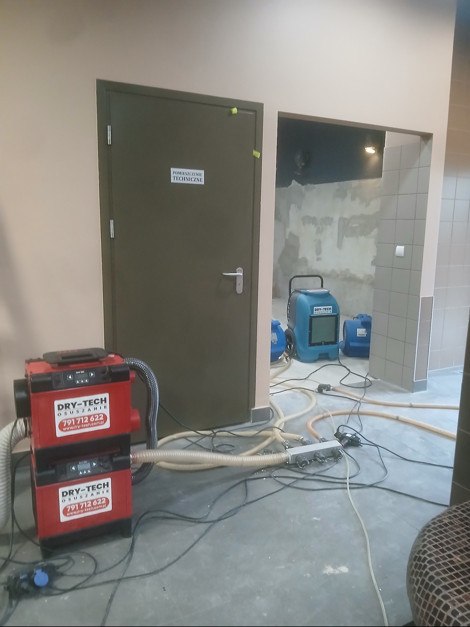 Osuszanie po zalaniu: czerwony i niebieski osuszacz DRY-TECH w pomieszczeniu z widocznymi śladami wilgoci na ścianach i podłodze, węże i kable rozłożone na posadzce.