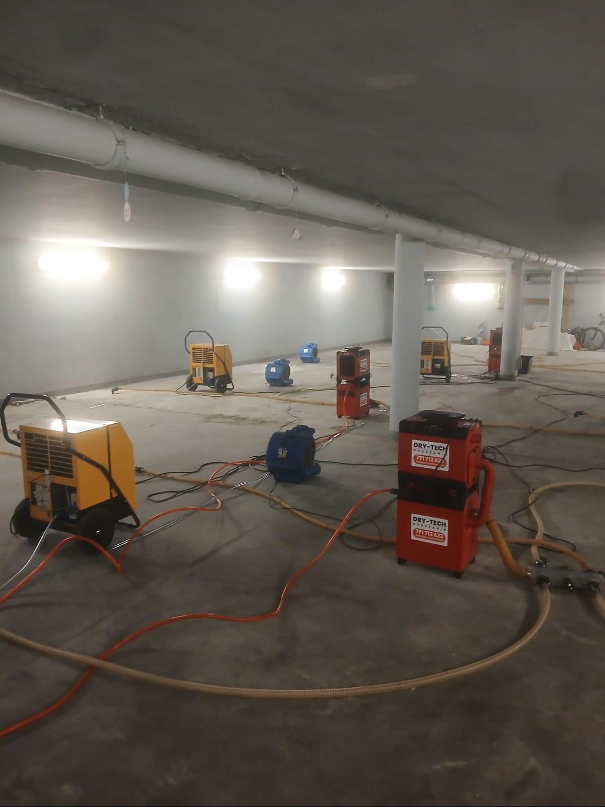 Rozstawione urządzenia do usuwania wilgoci w dużym, surowym pomieszczeniu z betonową podłogą i kolumnami. Widoczne żółte i czerwone nagrzewnice oraz niebieskie wentylatory.
