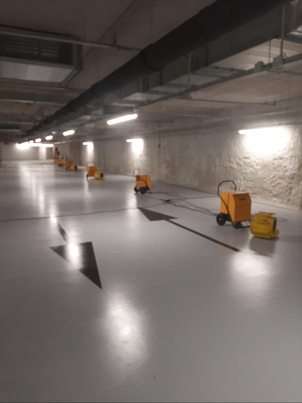 Przemysłowe osuszacze kondensacyjne na kółkach, ustawione w rzędzie wzdłuż betonowej posadzki z czarnymi strzałkami w hali garażowej. Widoczne przewody zasilające.