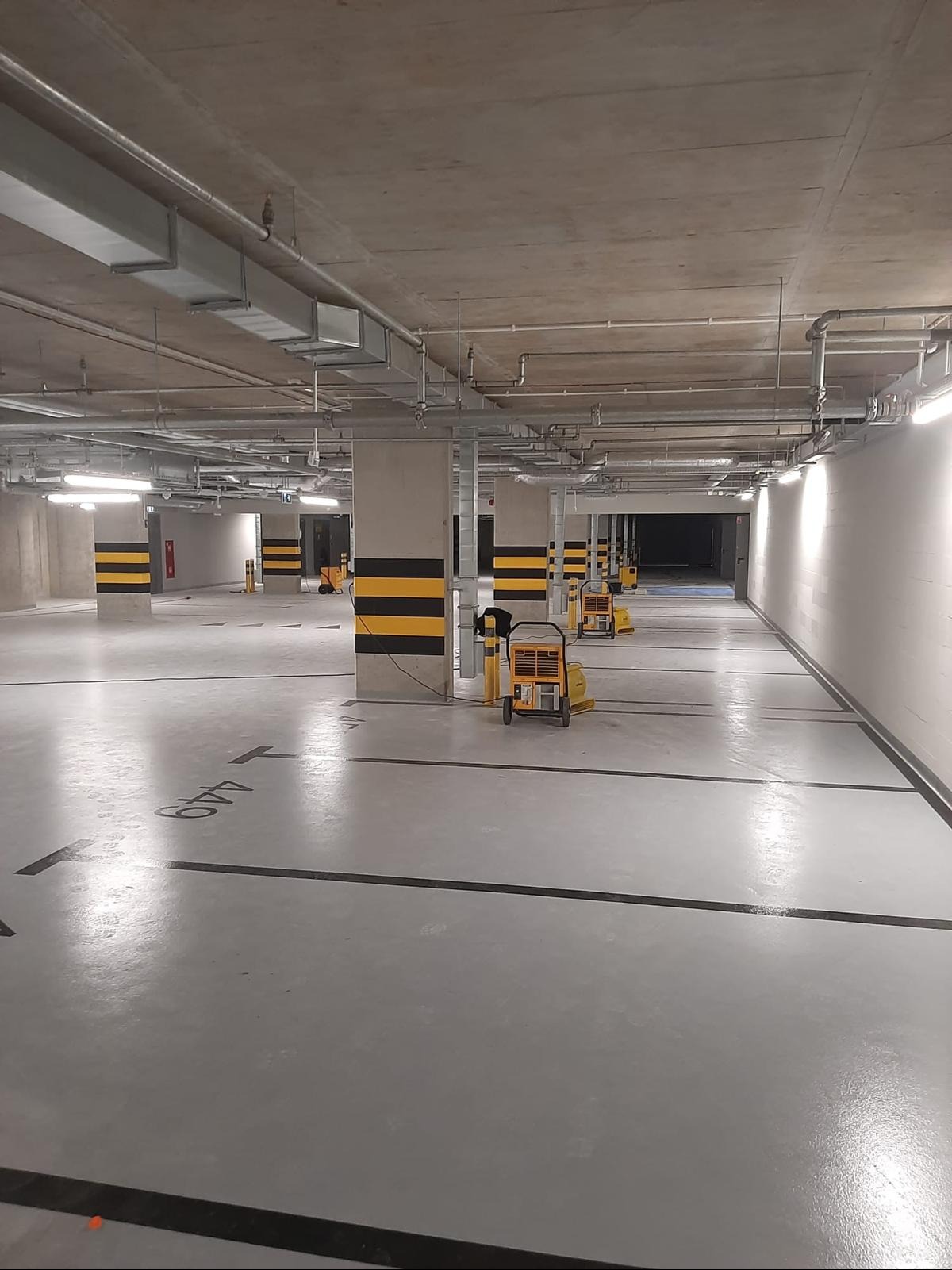 Osuszacze kondensacyjne na parkingu podziemnym po zalaniu. Widoczne słupy z oznaczeniami, instalacje pod sufitem i świeżo pomalowana posadzka.