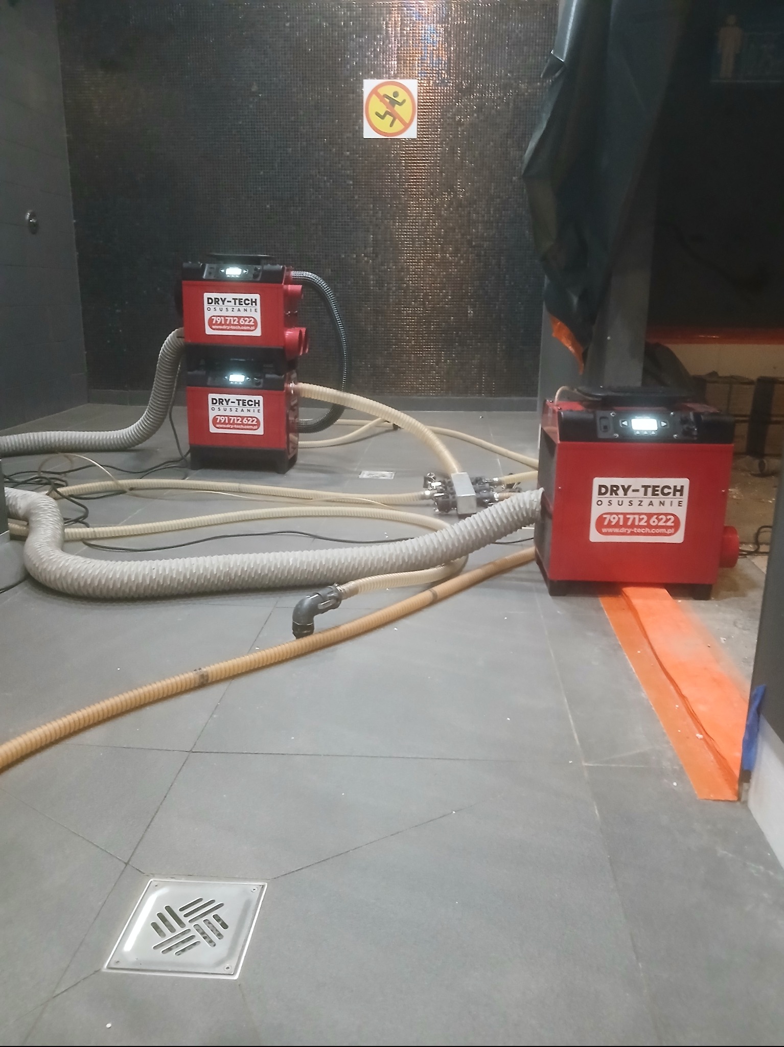 Dwa czerwone urządzenia z logiem Dry-Tech i rurami na szarej podłodze w pomieszczeniu z ciemną mozaiką na ścianie. Widoczny odpływ podłogowy i znak zakazu biegania.