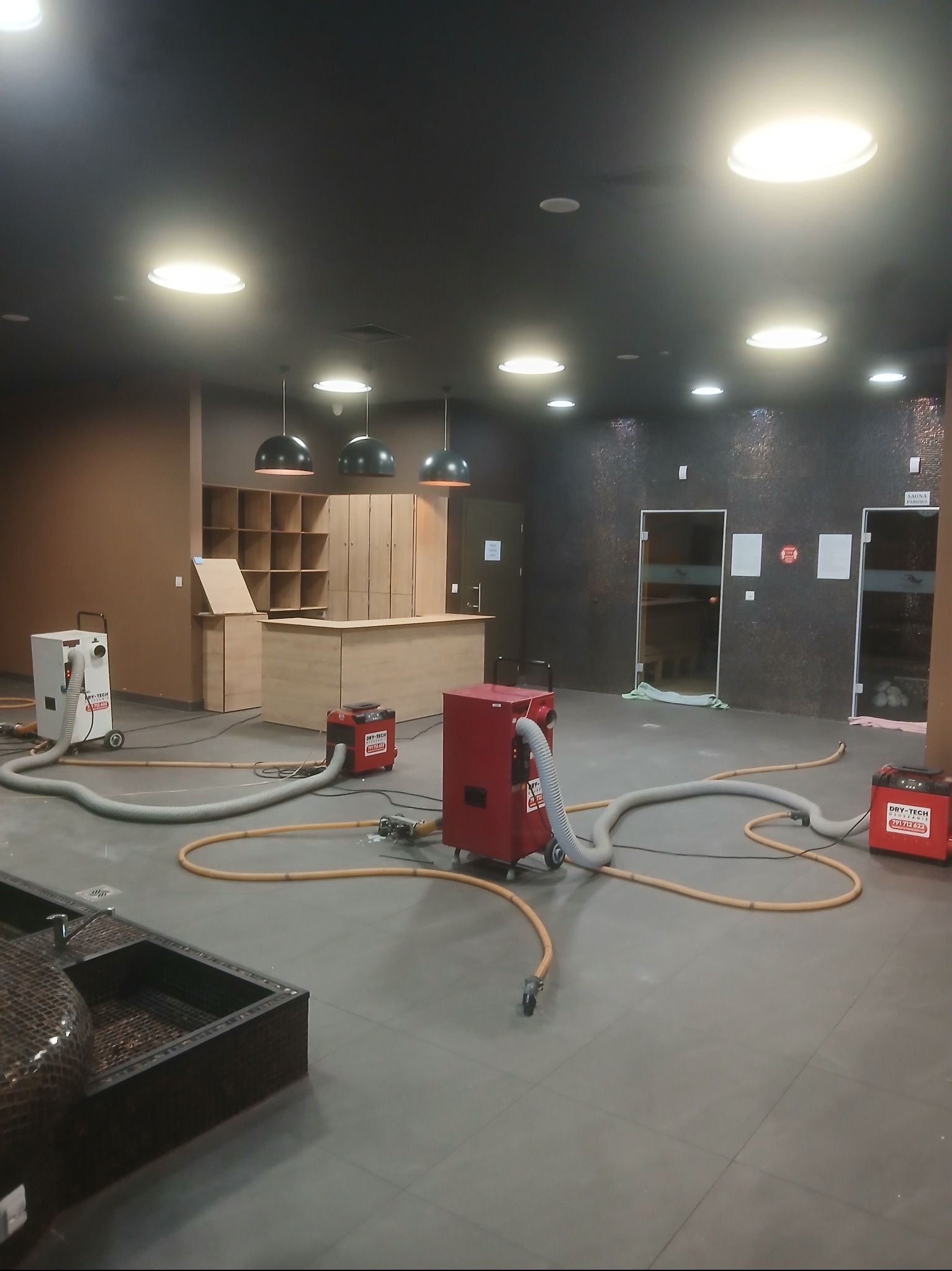 Wnętrze po zalaniu z rozstawionymi urządzeniami do usuwania wilgoci. Widoczne osuszacze i węże rozprowadzone po podłodze w pomieszczeniu z saunami i przebieralnią.
