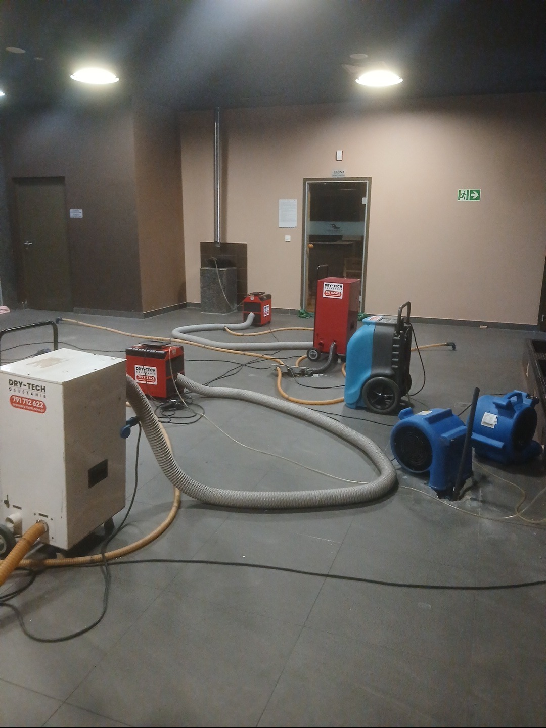 Osuszanie po zalaniu: kilka urządzeń Dry-Tech z podłączonymi wężami, wentylatory, szara podłoga, beżowe ściany, widoczne drzwi i wyjście ewakuacyjne.