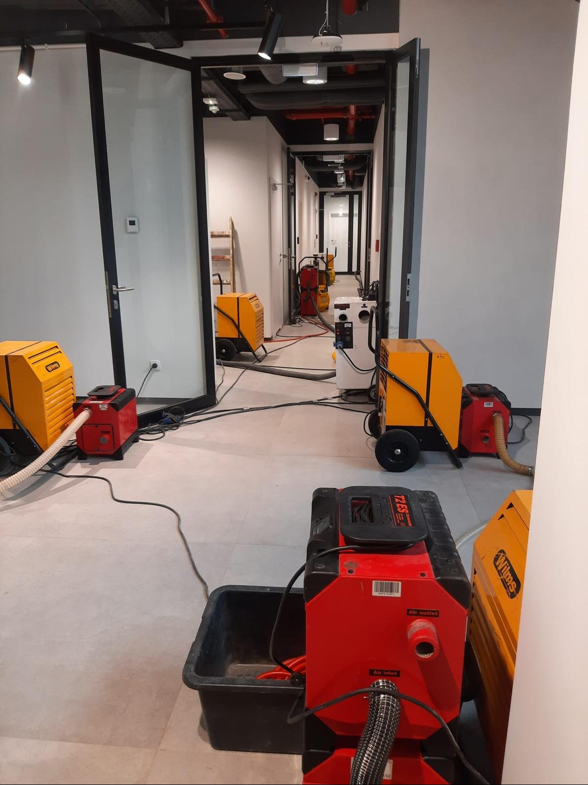 Korytarz biurowy z rozstawionymi, pracującymi osuszaczami powietrza Wilms w kolorach żółto-czerwonym. Widoczne przewody i detale urządzeń. Perspektywa w głąb korytarza.