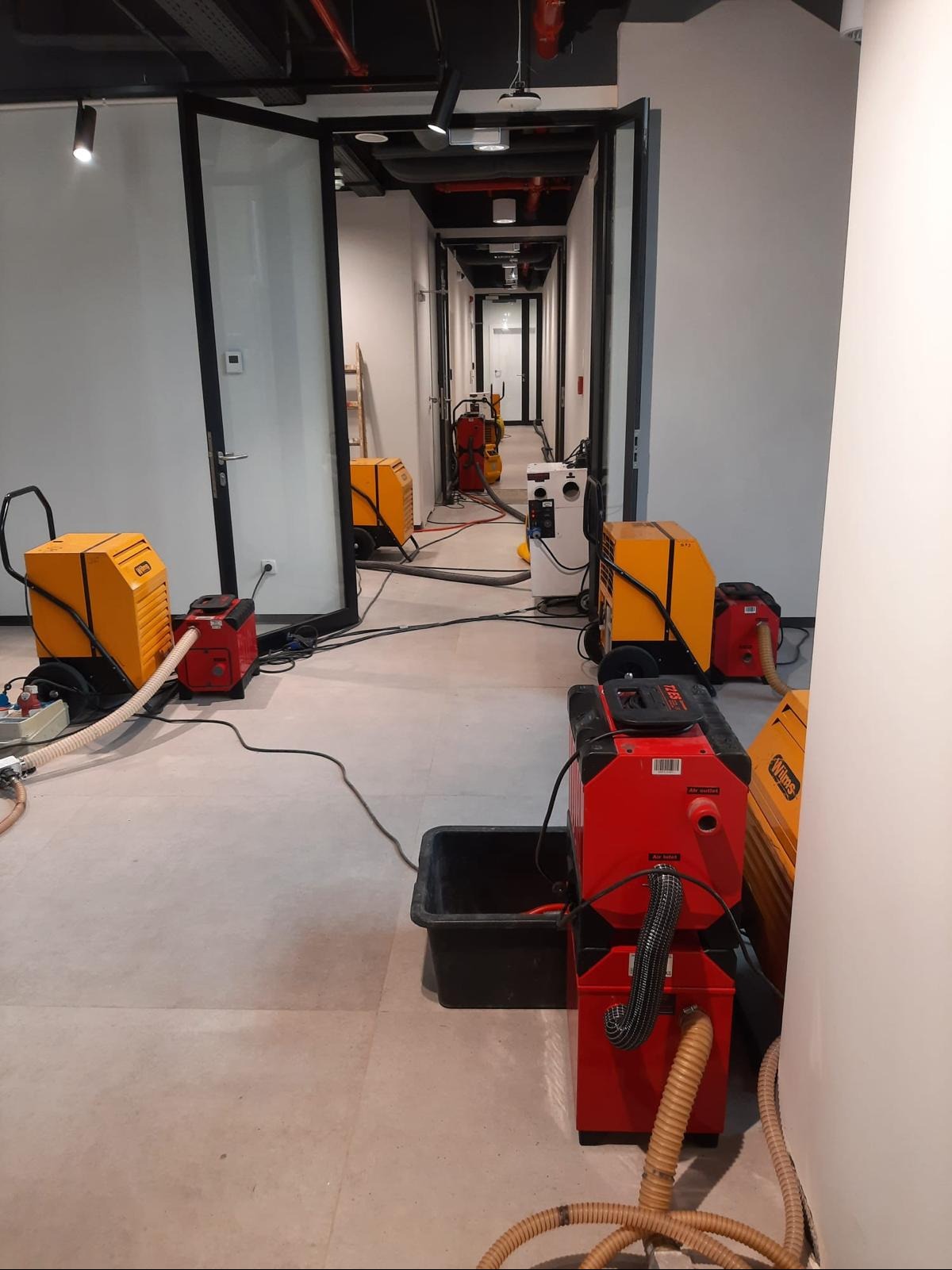Korytarz biurowy z rozstawionymi, pracującymi urządzeniami w kolorze żółtym i czerwonym. Czarny pojemnik na wodę. Widoczne kable i węże. Etap prac.
