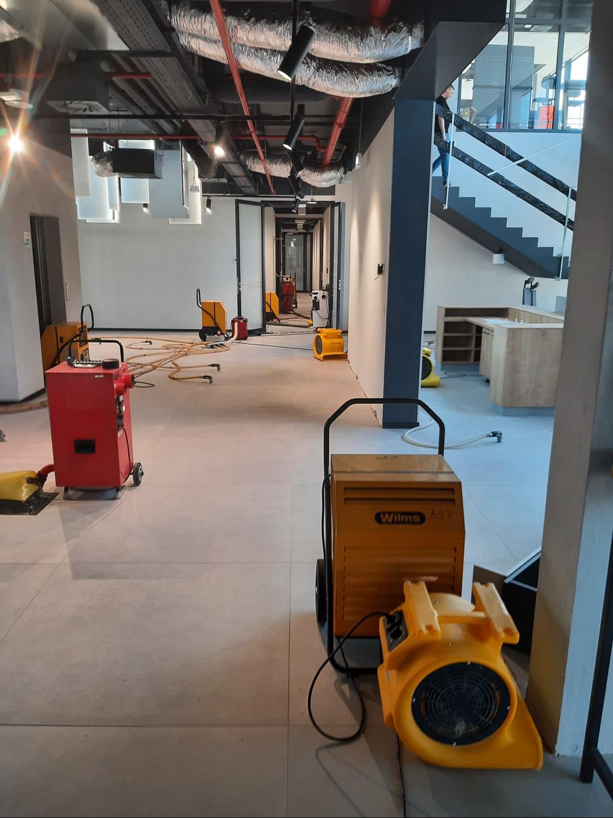 Profesjonalne osuszanie biura: widok na rozstawione osuszacze powietrza Wilms 627 i inne urządzenia, kable i węże na jasnej podłodze w przestronnym wnętrzu biurowym.