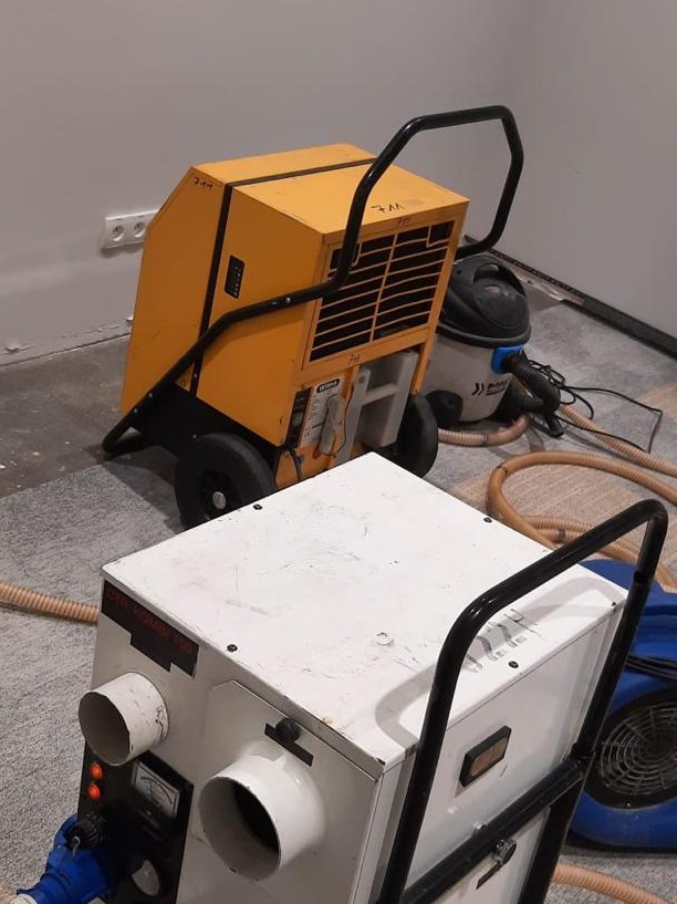 Dwa przemysłowe urządzenia do osuszania, żółte i białe, z czarnymi uchwytami, stoją na szarej podłodze w pomieszczeniu. W tle widoczny odkurzacz i fragment ściany.