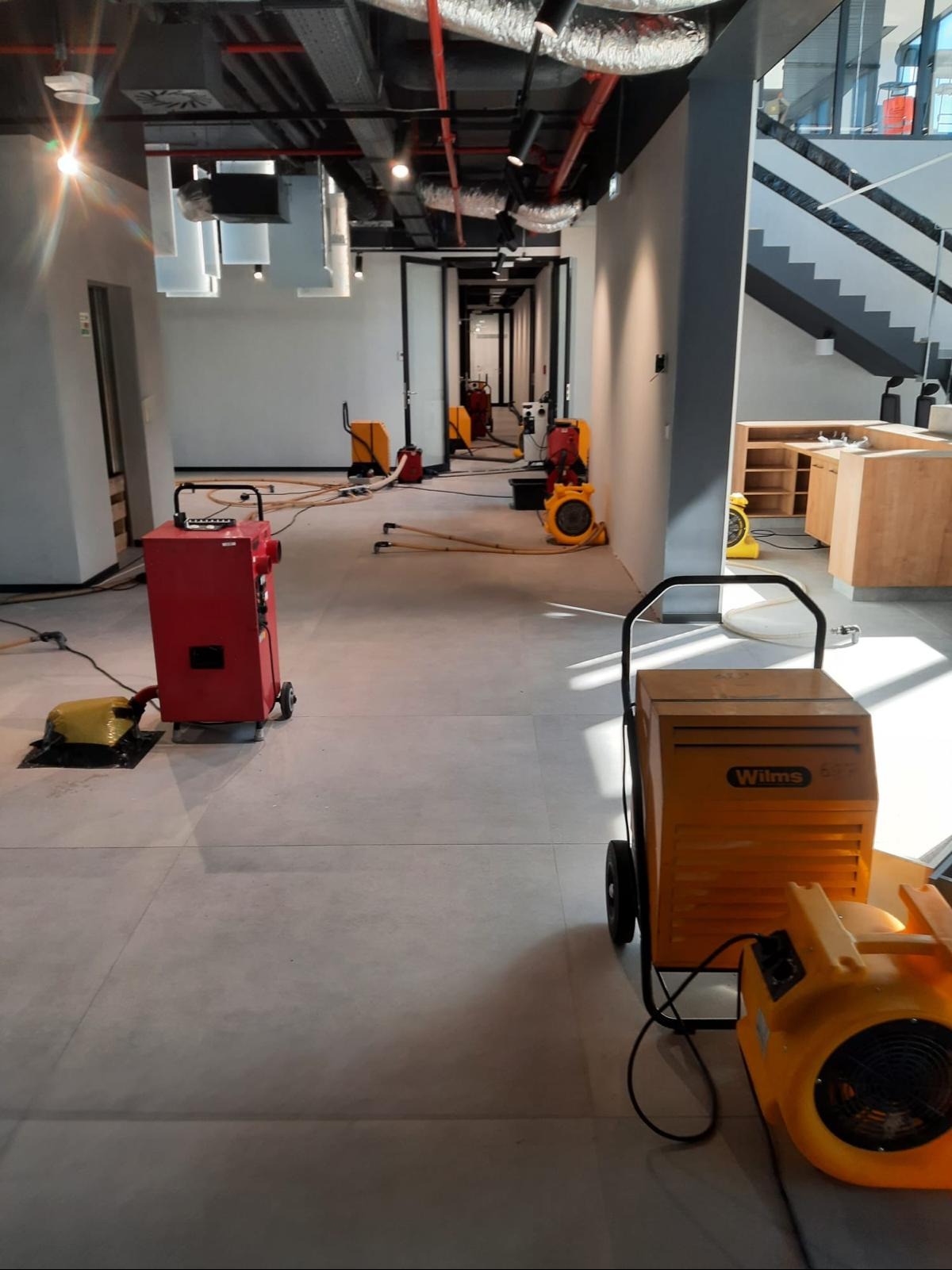 Sprzęt do usuwania wilgoci w nowoczesnym biurze: czerwony i żółty, z przewodami na szarej podłodze. Widoczne elementy instalacji pod sufitem.