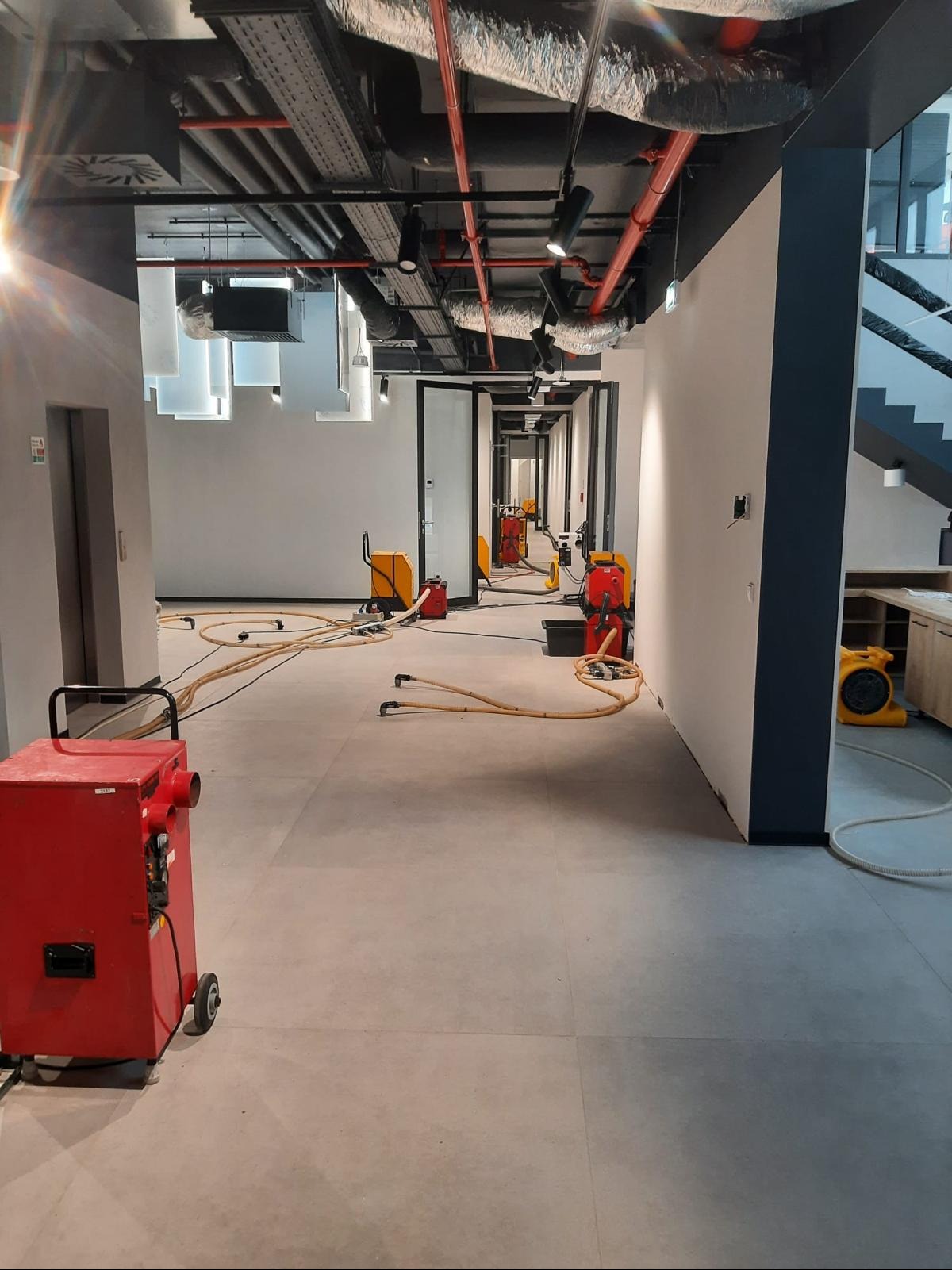 Korytarz biurowy w trakcie osuszania po zalaniu. Widoczne profesjonalne osuszacze powietrza i węże rozprowadzające powietrze. Elementy instalacji pod sufitem.