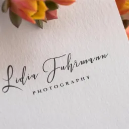 Eleganckie logo 'Lidia Fuhrmann Photography' w czerni na teksturowanym, kremowym papierze, ozdobione kwiatami w odcieniach pomarańczy i żółci, z delikatnym cieniem i widocznym znakiem wodnym...