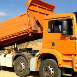 Pomarańczowa wywrotka MAN z podniesioną skrzynią ładunkową, widoczna na piaszczystym terenie w słoneczny dzień.