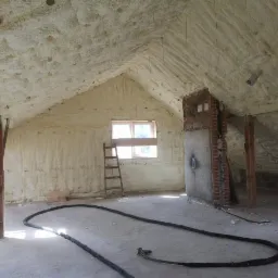 Poddasze po ociepleniu pianką: ściany, sufit i skosy pokryte jasną pianą, widoczne drewniane słupy, okno z drabiną i przewód na podłodze.