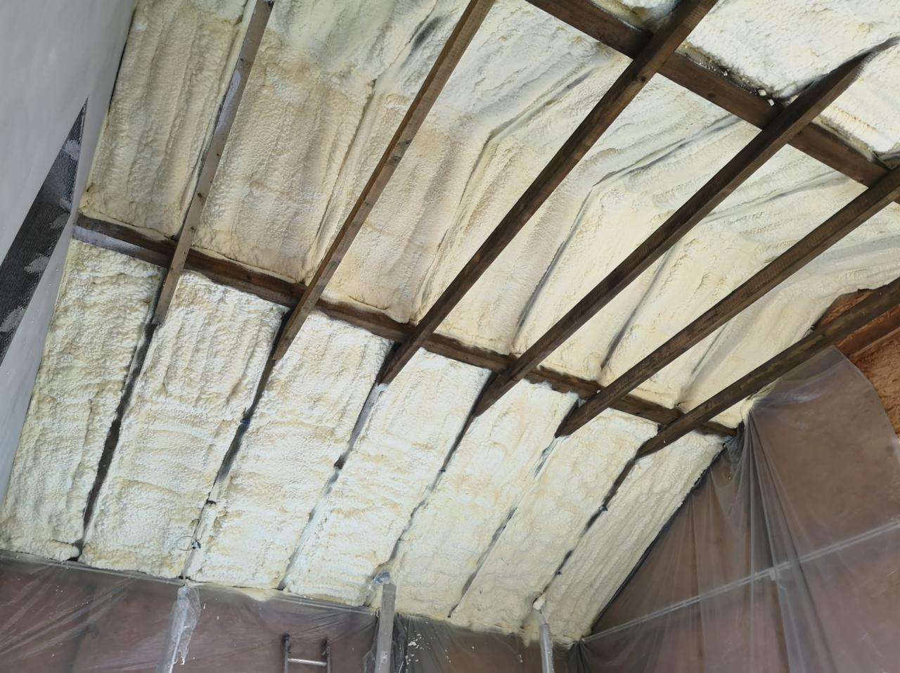 Poddasze z drewnianą konstrukcją pokryte grubą warstwą jasnej pianki izolacyjnej, widoczne folie ochronne i drabina w tle.